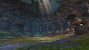 A screenshot of the domed arena, from the right of the entrance. The large, empty combat area can be seen with red and blue torches and drake statues around the edge. The arena is surrounded by stairs to an upper level.
